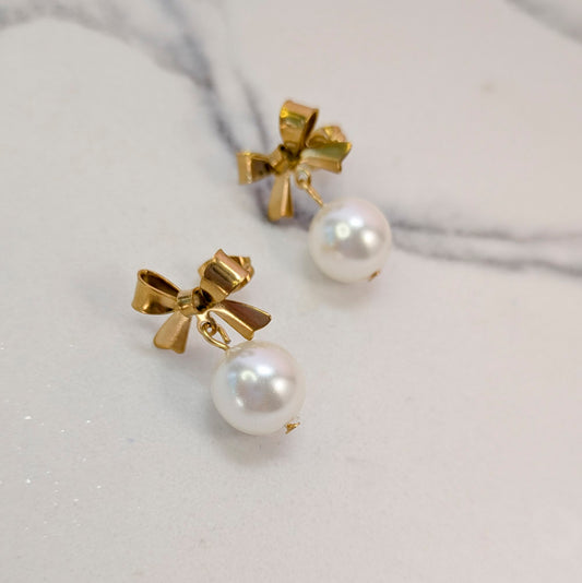 A pair of gold bow stud earrings with white pearl drops, displayed on a light marble surface.