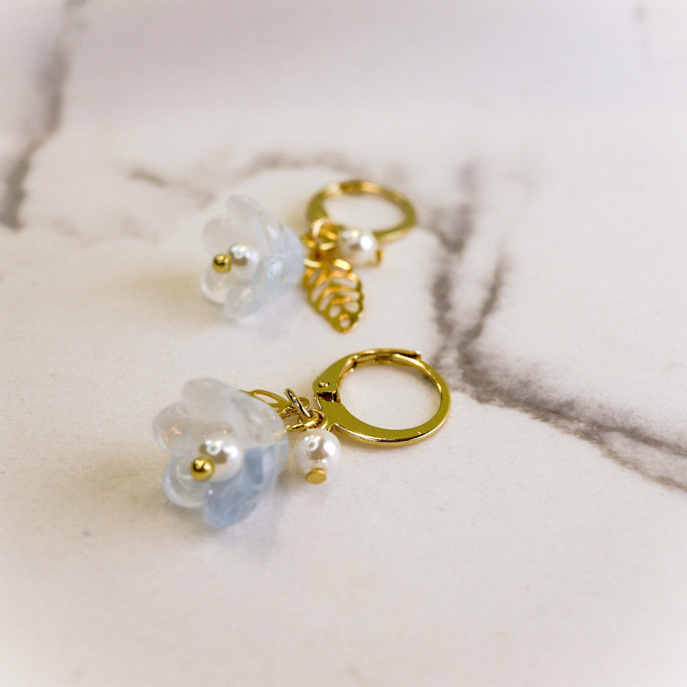 A pair of gold-tone hoop earrings with translucent pale blue flower charms, small white pearl beads, and a gold leaf charm, displayed on a light marble surface.