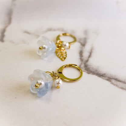 A pair of gold-tone hoop earrings with translucent pale blue flower charms, small white pearl beads, and a gold leaf charm, displayed on a light marble surface.