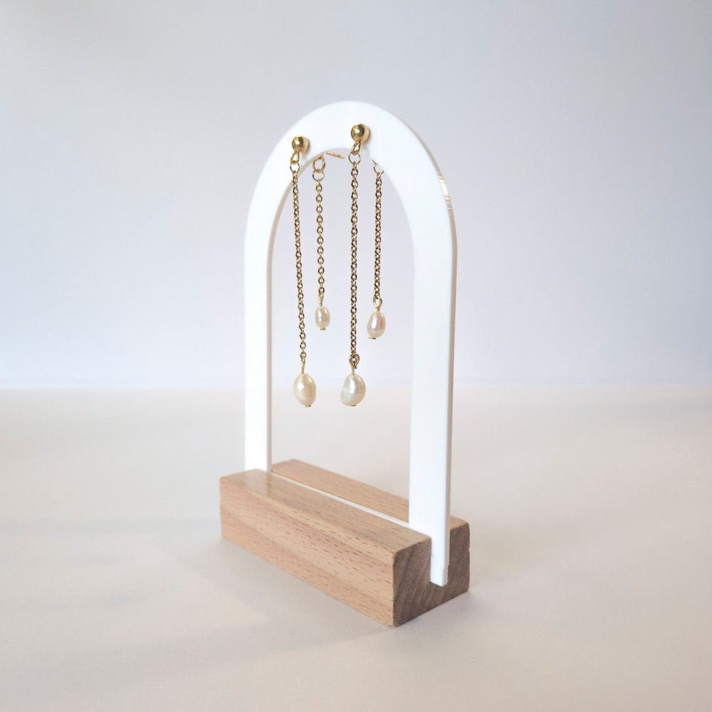 A pair of gold chain earrings with white pearl drops displayed on a white and light wood stand against a clean white background.