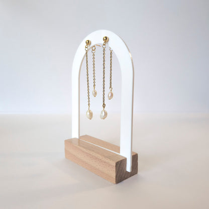 A pair of gold chain earrings with white pearl drops displayed on a white and light wood stand against a clean white background.