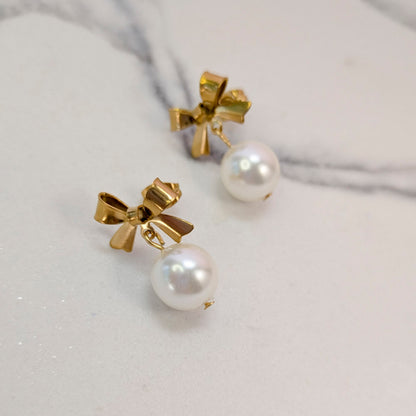 A pair of gold bow stud earrings with white pearl drops, displayed on a light marble surface.