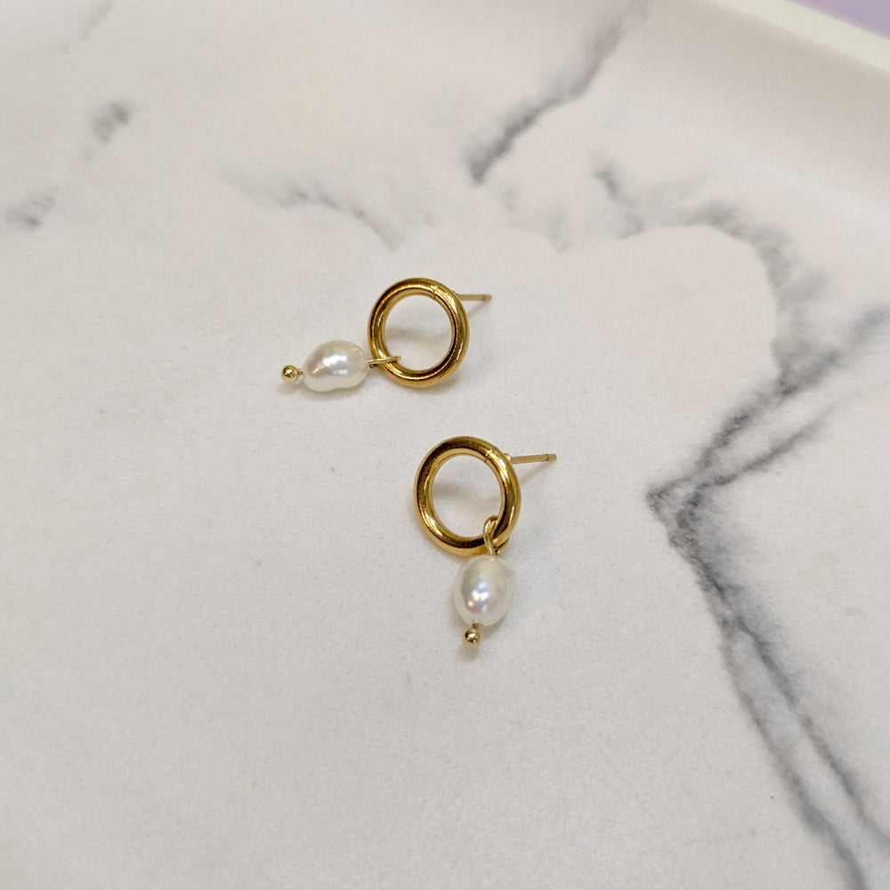 A pair of gold hoop earrings with a small white baroque pearl dangling below, displayed on a white marble surface.