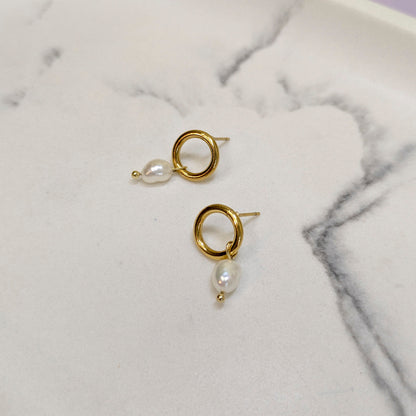 A pair of gold hoop earrings with a small white baroque pearl dangling below, displayed on a white marble surface.