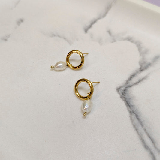 A pair of gold hoop earrings with a small white baroque pearl dangling below, displayed on a white marble surface.